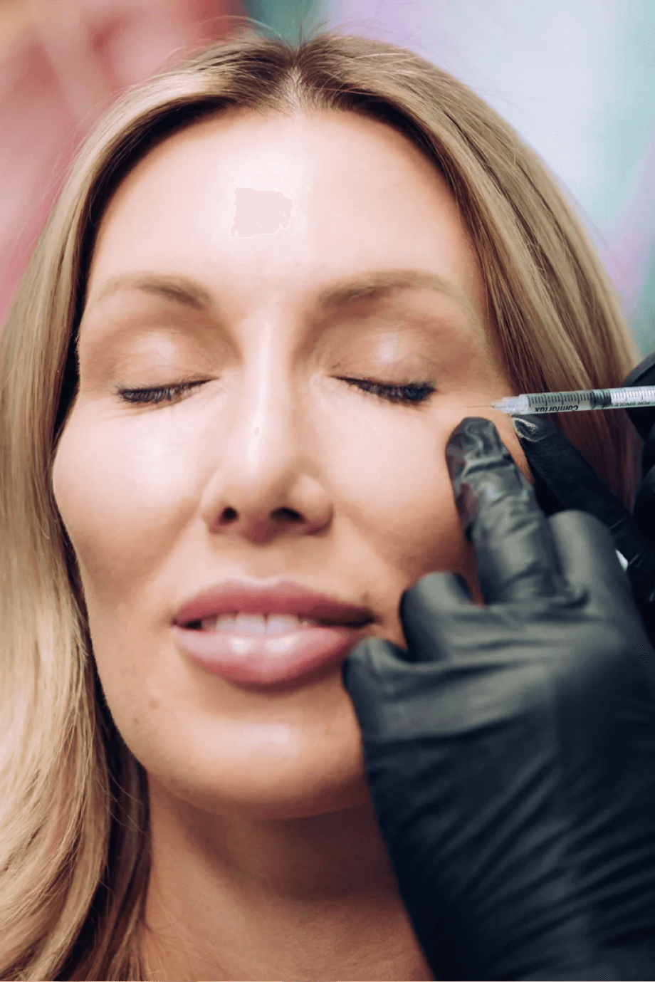 A close-up of a woman receiving a facial injection near her eye at a wellness clinic. She appears relaxed with her eyes closed, while a professional in black gloves administers the injection.