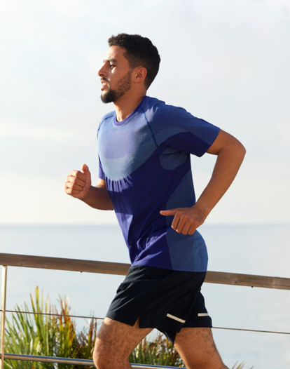 A man jogging outdoors near the ocean, wearing a blue athletic shirt and black shorts.