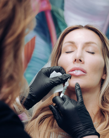 A woman receiving a lip filler injection from a professional wearing black gloves, with her eyes closed