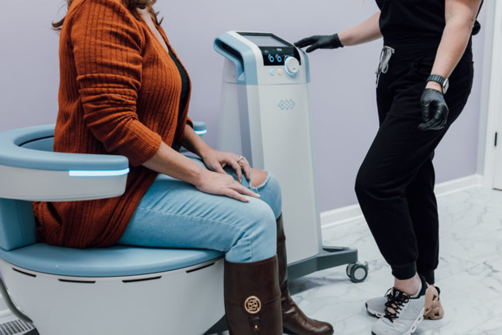 A woman sitting on a wellness treatment chair while a technician prepares to operate the machine.