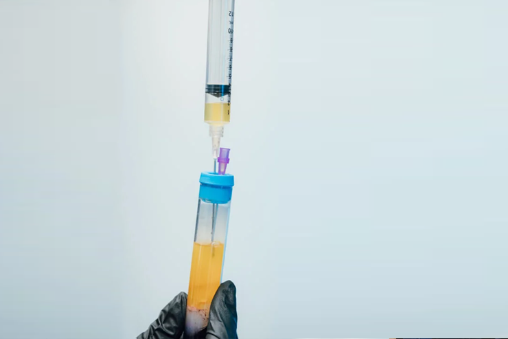 A gloved hand holds a test tube with a yellow liquid while a syringe filled with a yellow liquid is being inserted into the test tube.