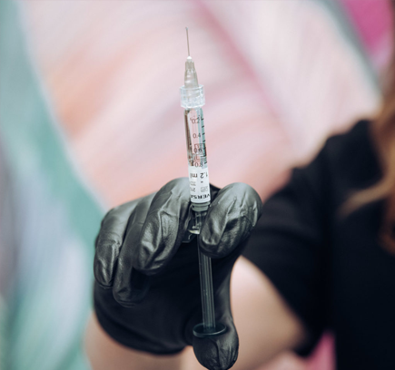 A gloved hand holds a syringe filled with a clear liquid. The syringe is positioned vertically, with the needle pointing upwards. The background is blurry and appears to be a colorful wall.